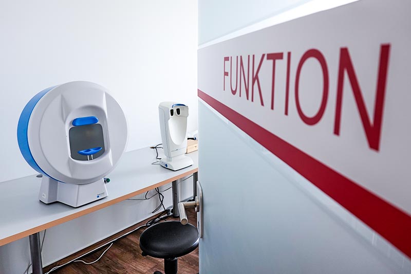 Ein medizinisches Gerät steht auf einem Tisch neben einem Stuhl, während im Hintergrund eine Wand mit der Aufschrift "FUNKTION" zu sehen ist.