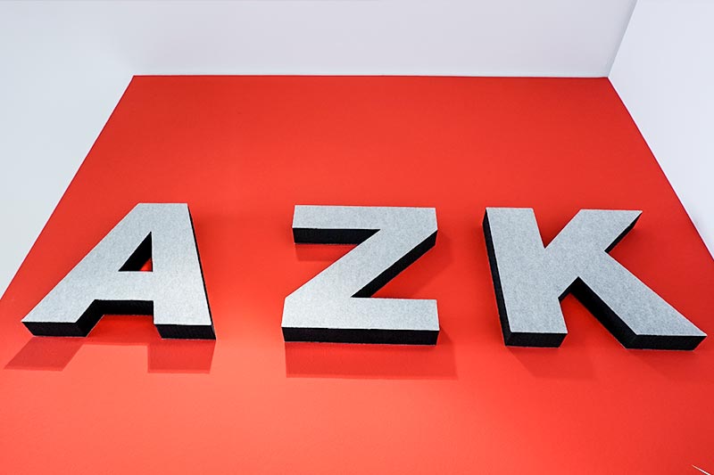 Drei silberne 3D-Buchstaben "AZK" auf einem roten Hintergrund.